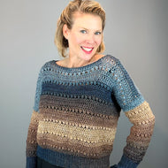 A smiling woman wearing a crocheted long-sleeve sweater with horizontal stripes in blue, brown, and beige, posed against a solid gray studio background.