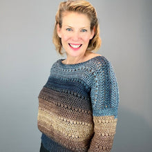 Load image into Gallery viewer, A smiling woman with blonde hair wearing a hand-crocheted long-sleeve sweater with a blue, brown, and beige color-blocked pattern, set against a plain gray studio background.
